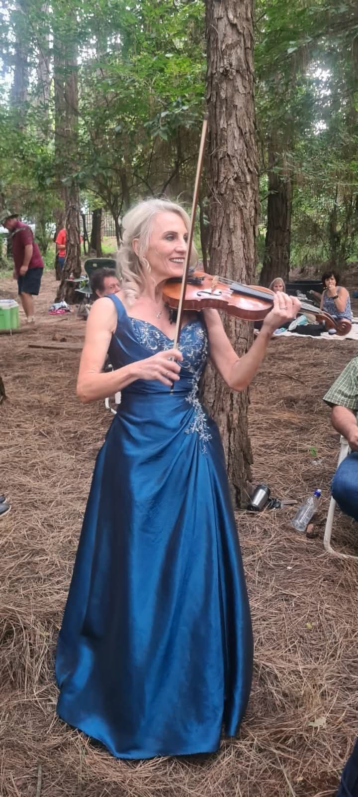 Esmé Bergh in a long blue gown playing the violin in a pine forest, with people seated among the trees