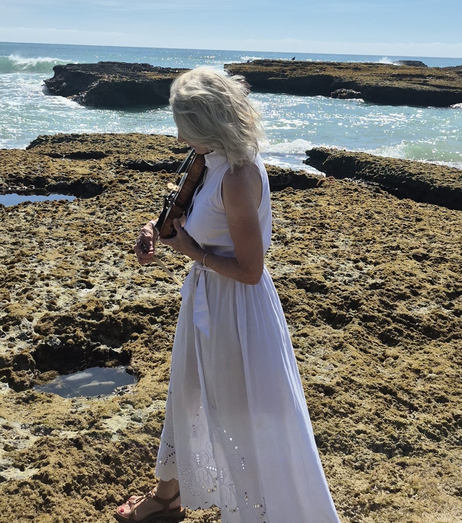Esmé Bergh playing the violin in a white dress on a rocky coastal shelf with the sea behind her