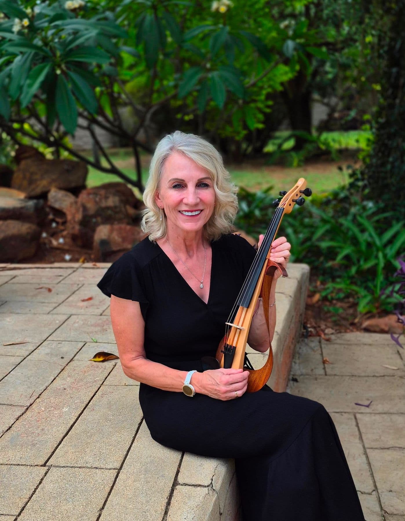 Esmé Bergh seated on a low stone wall in a garden, smiling, holding an electric violin with an open wooden frame