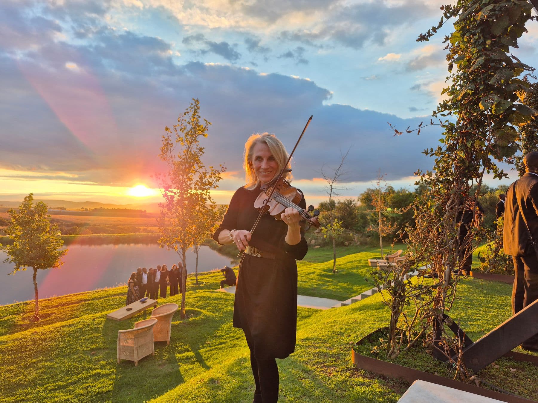 Esmé Bergh playing the violin on a lawn at sunset, with people gathered beside a lake below her