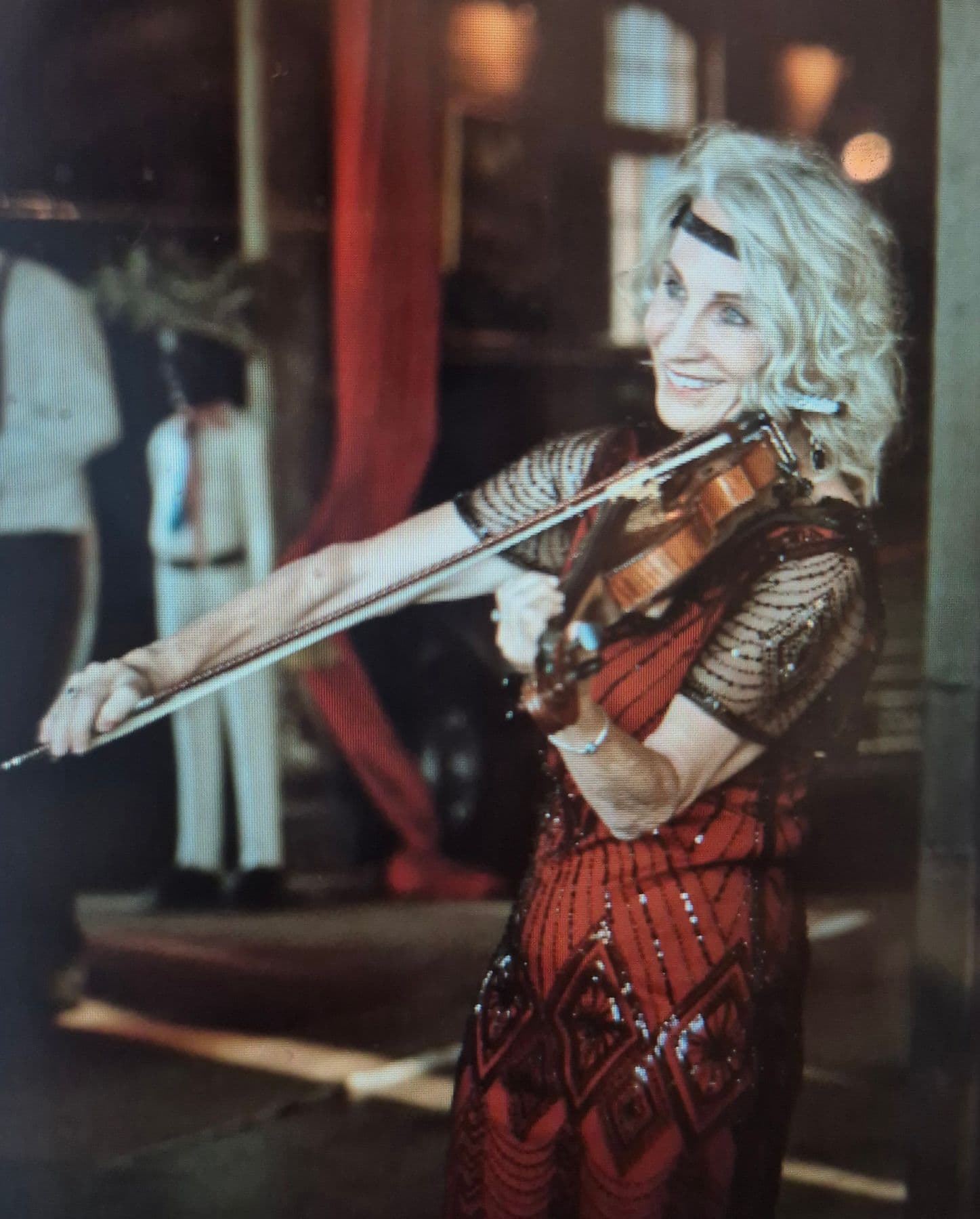 Esmé Bergh playing the violin indoors in a red beaded dress and a headband, with people behind her