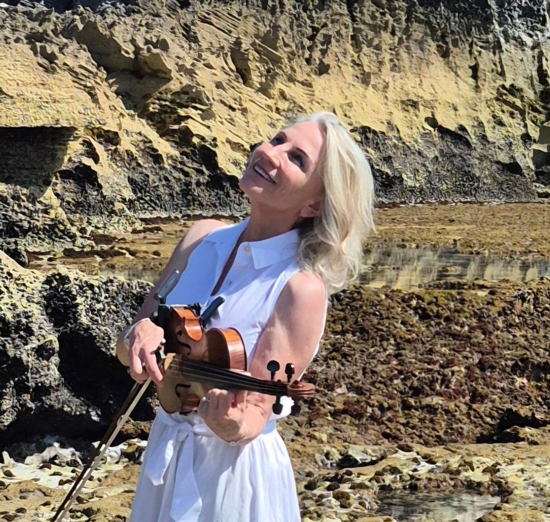 Esmé Bergh in a white dress on a rocky shore beside rock pools, smiling up at the sky with her violin