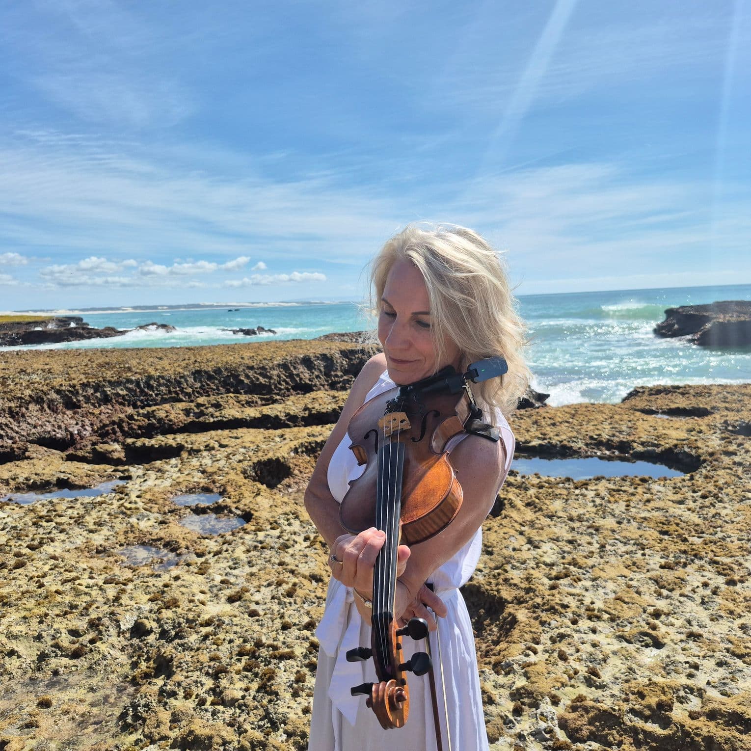 Esmé Bergh standing on a rocky shore holding her violin, the open sea behind her