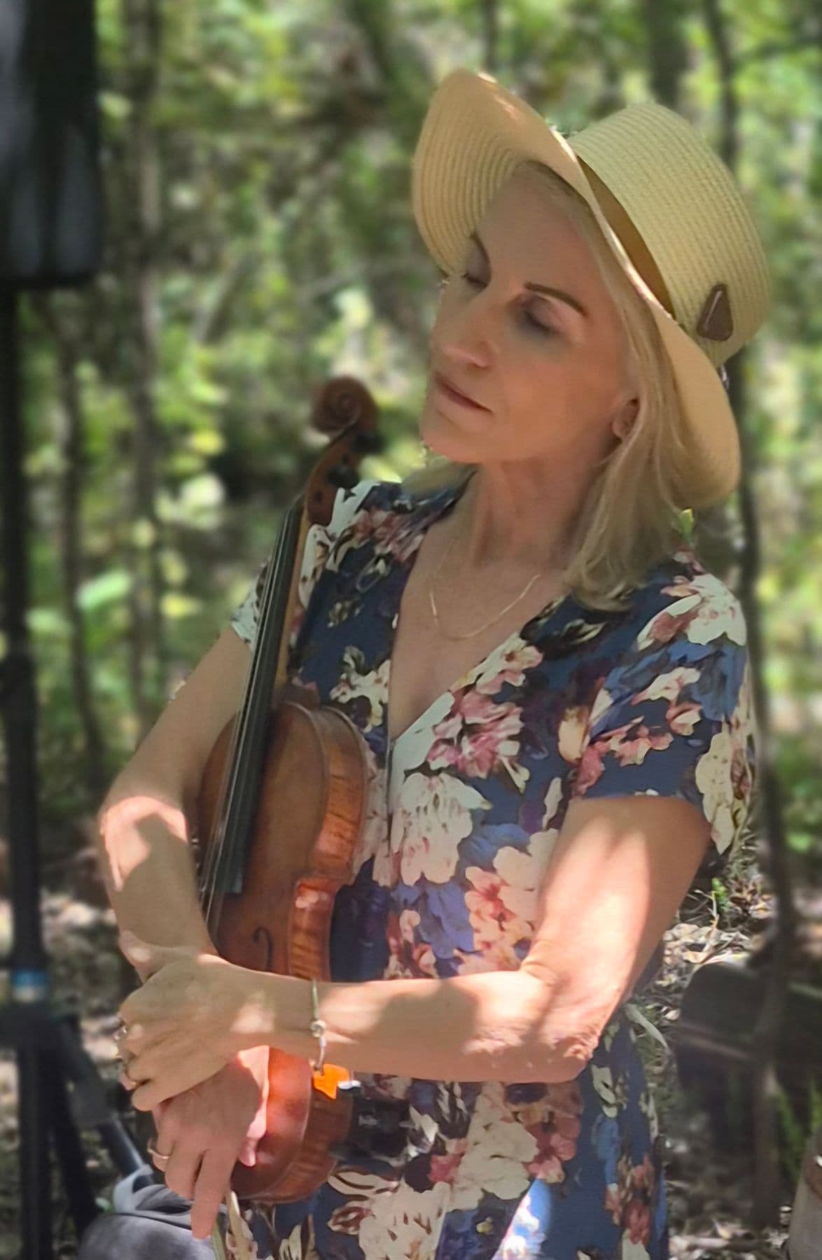 Esmé Bergh in a straw hat and a floral dress, eyes closed, holding her violin among trees