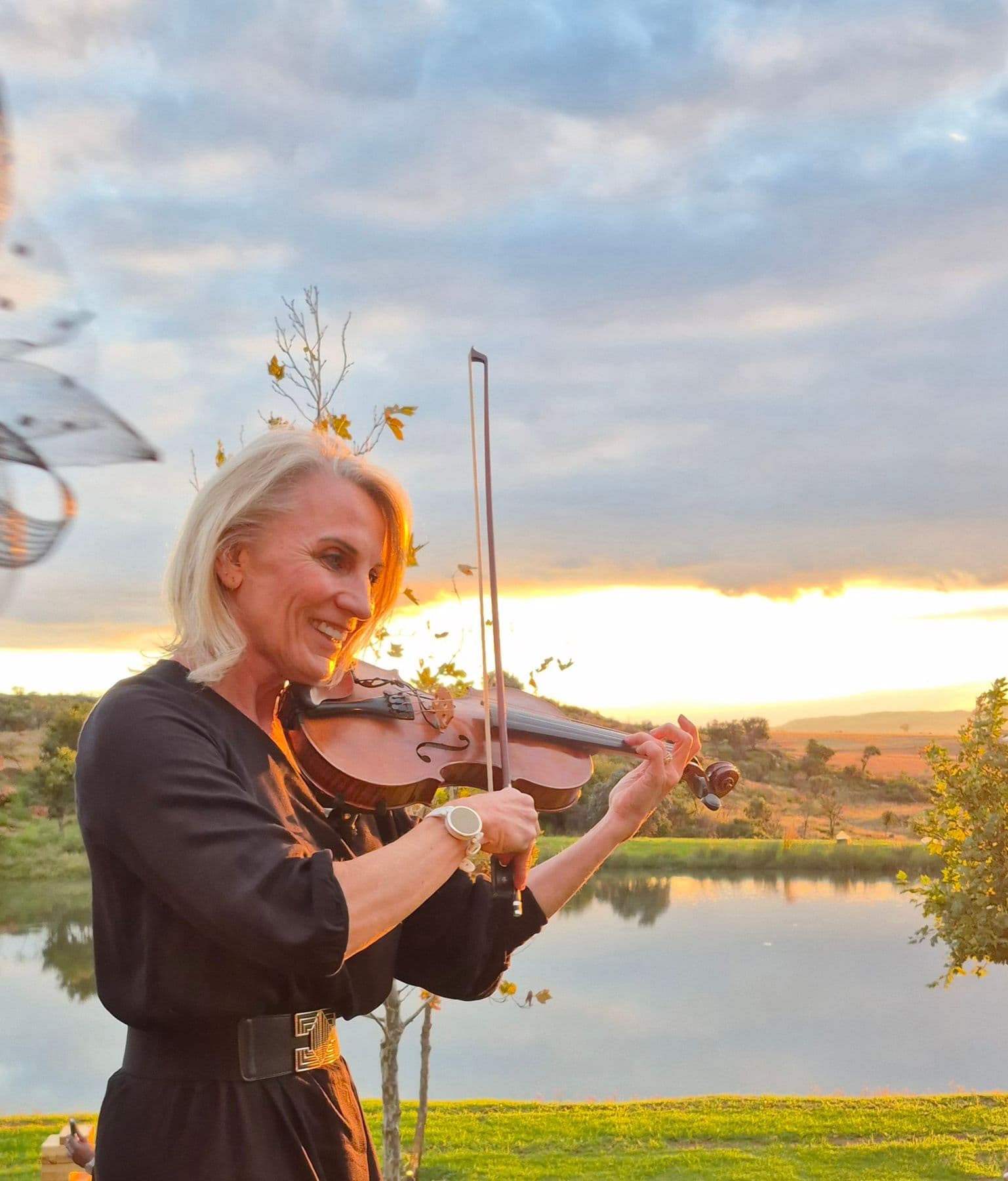 Esmé Bergh smiling as she plays the violin at sunset, a lake and open farmland behind her