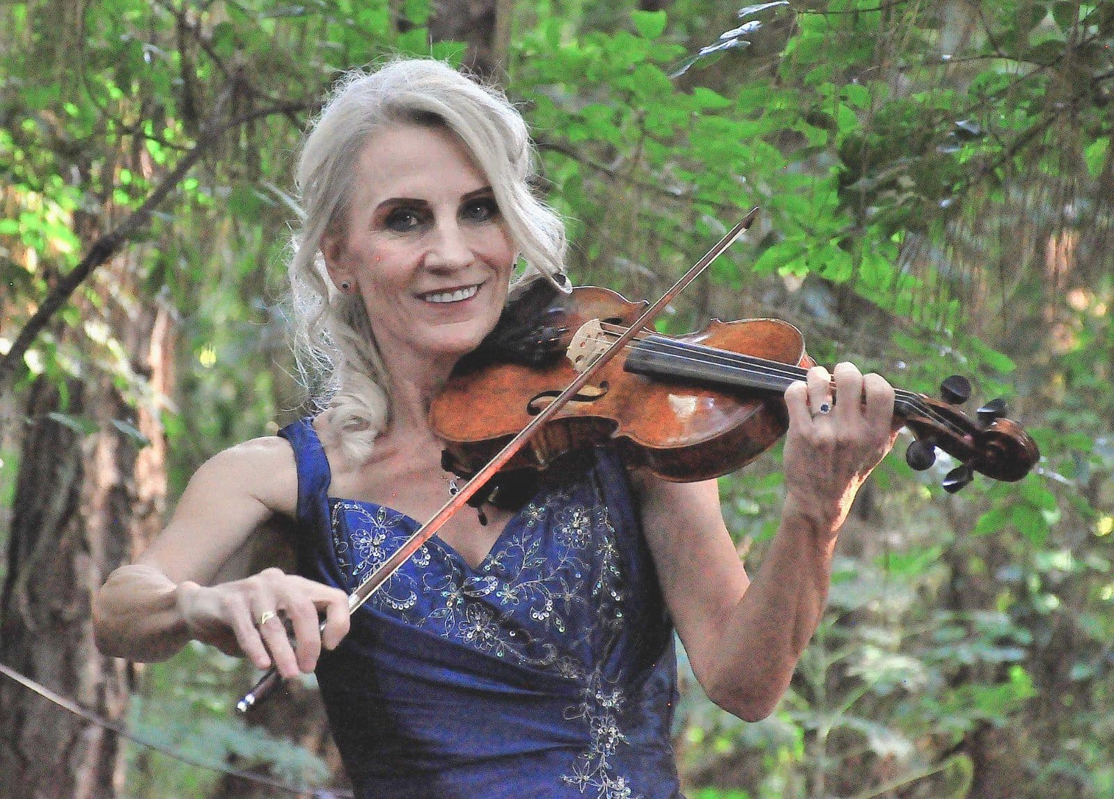 Esmé Bergh playing the violin outdoors among trees, in a deep blue beaded dress