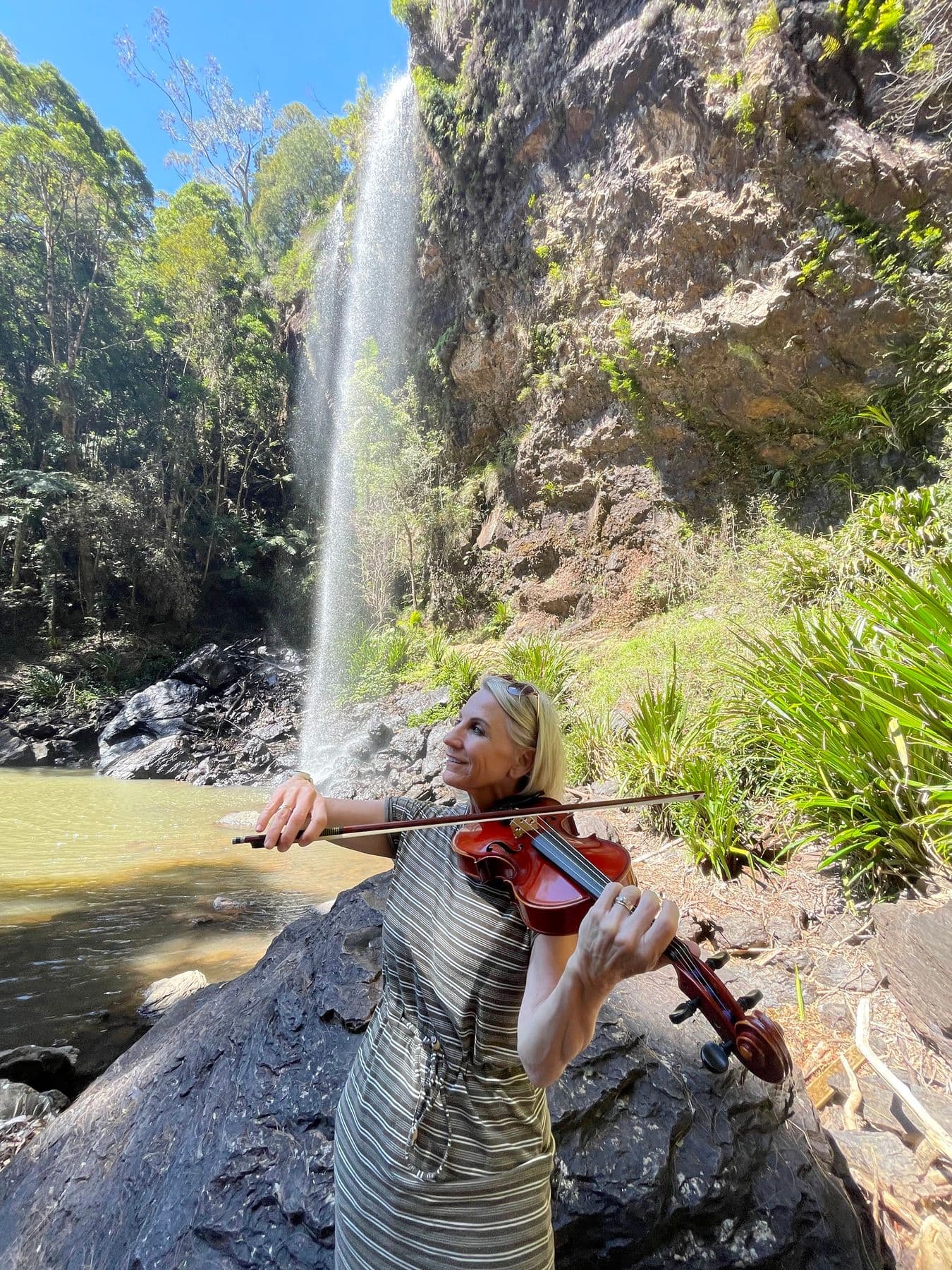 Esmé Bergh playing the violin on a boulder beside a tall waterfall and a forest pool